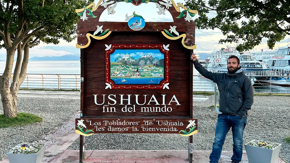 Uomo di fianco al cartello di ingresso nella città di Ushuaia